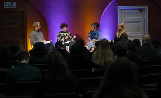 Four people sit on a stage with microphones, against colourful lighting and branding that says Shameless! Festival