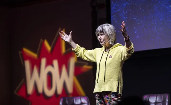 Jude (a white woman in a yellow top) stands on a stage with her hands in the air