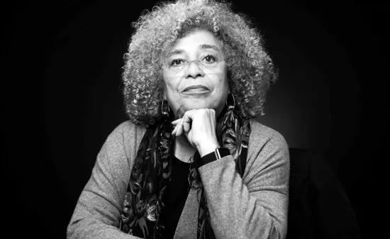 A black and white headshot of Angela Davis, a woman with grey afro hair and glasses, she is sat down with her hand on her chin.